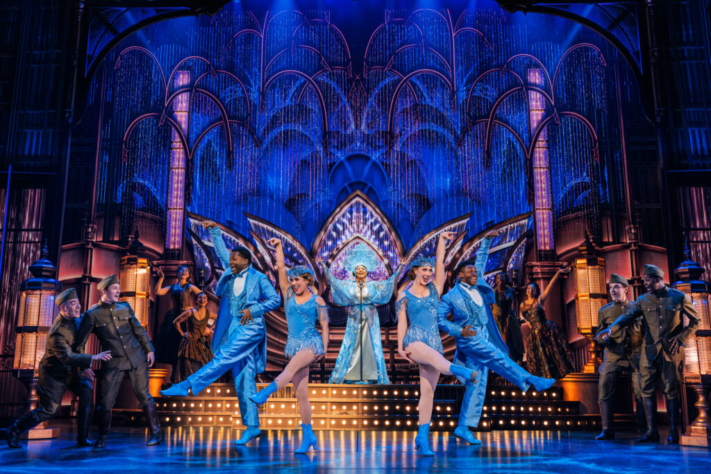 Noah J. Ricketts stands center stage in the Broadway production of The Great Gatsby, illuminated by a spotlight against a striking, blue-lit backdrop, with silhouetted cast members striking elegant poses in the background. Photo by Matthew Murphy and Evan Zimmerman.