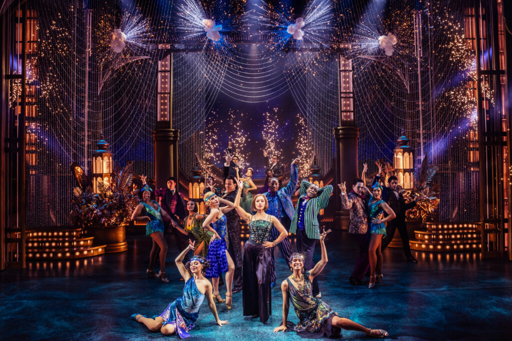 Samantha Pauly (center) and the cast of the Broadway production of The Great Gatsby perform onstage under a dazzling backdrop of cascading lights, golden decor, and fireworks, with a glamorous 1920s-inspired setting. Photo by Matthew Murphy and Evan Zimmerman.