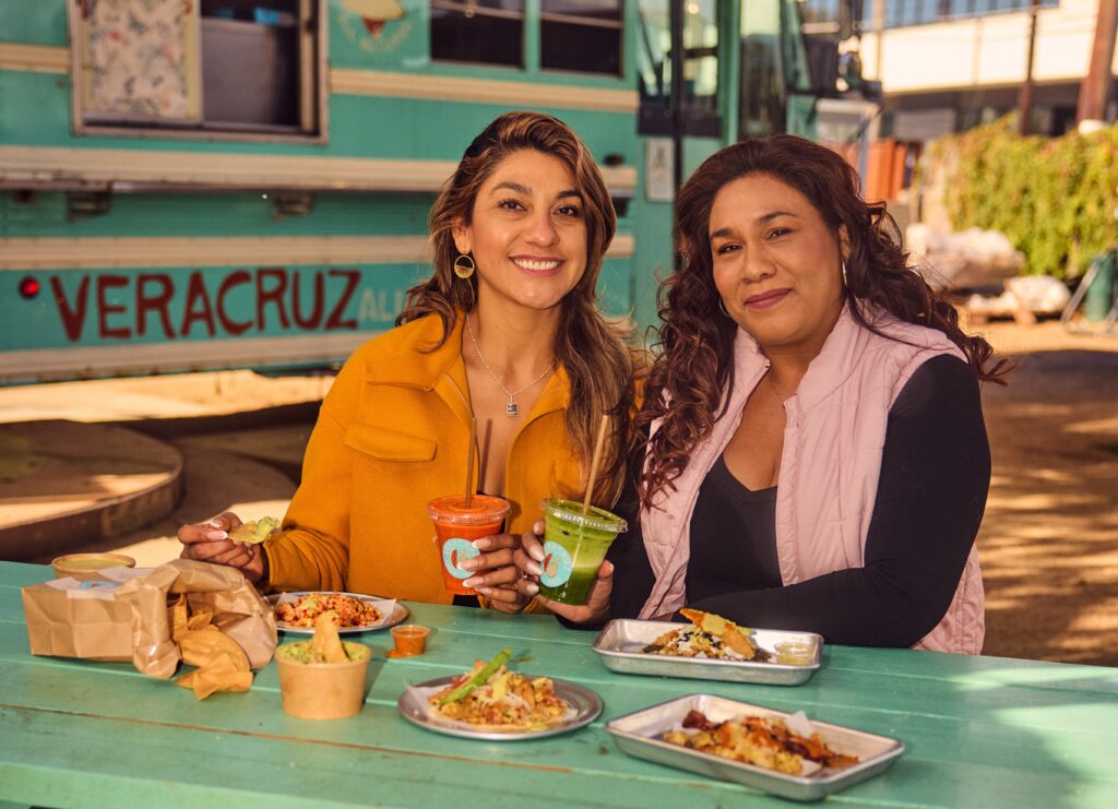 Sister chefs sitting in front of their food truck in Austin, Texas with several tacos.