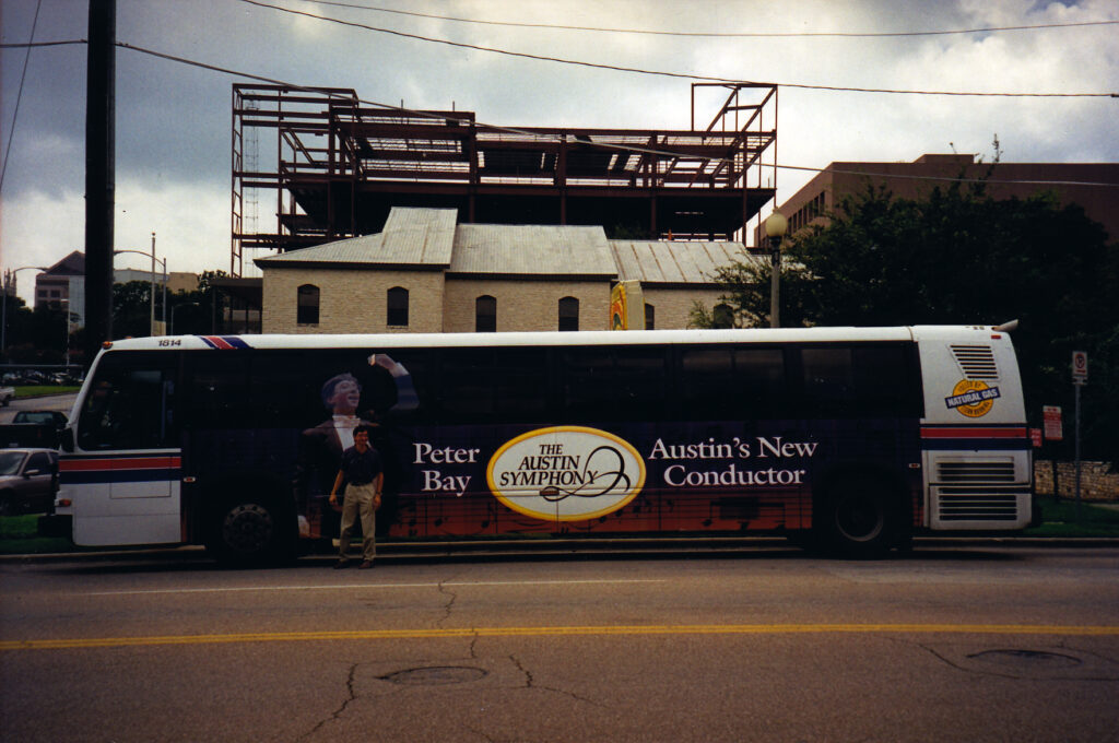 Peter Bay stands in front of a city bus displaying an advertisement announcing him as the Austin Symphony Orchestra’s new music director.